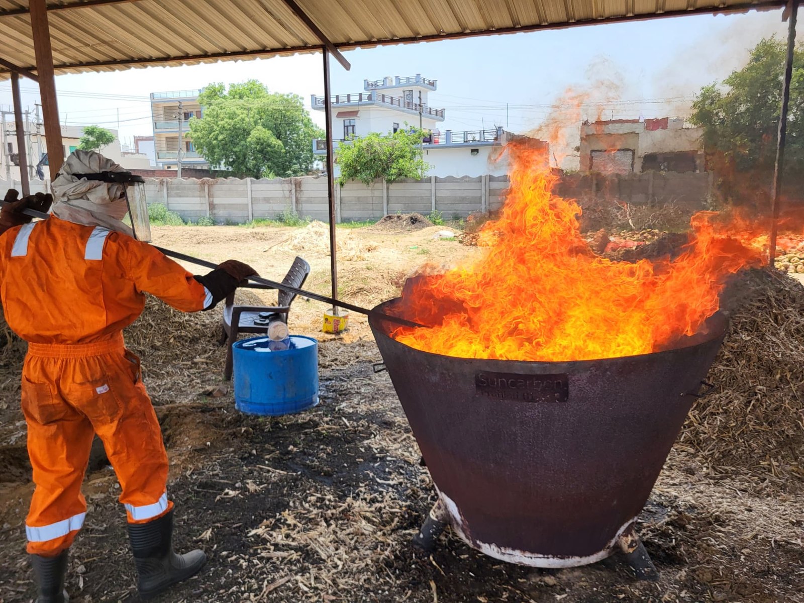 Operator in protective clothing managing a biochar kiln flame at a distributed production site in India